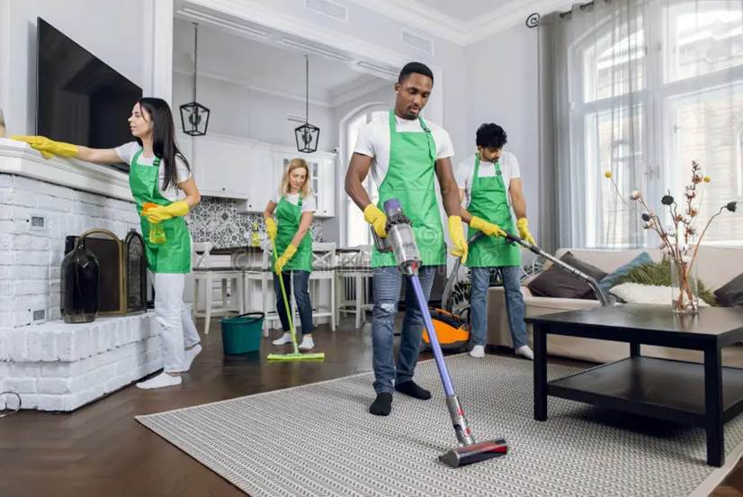 Four professional cleaners wearing matching green aprons and yellow gloves thoroughly cleaning a modern, spacious living room and kitchen area.