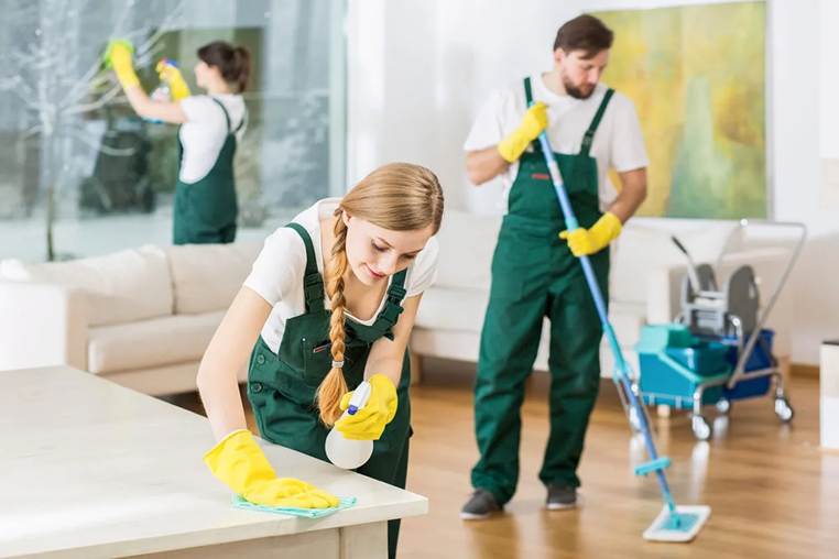 Three professional cleaners in green overalls and yellow gloves cleaning a bright, modern living room.